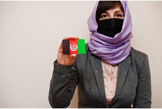 Portrait of young muslim woman wearing formal wear, protect face mask and hijab head scarf, hold Afghanistan flag card against isolated background. Coronavirus country concept.